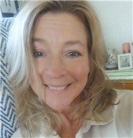 En leende kvinna med blont hår och blus, fotograferad nära en ljus bakgrund.